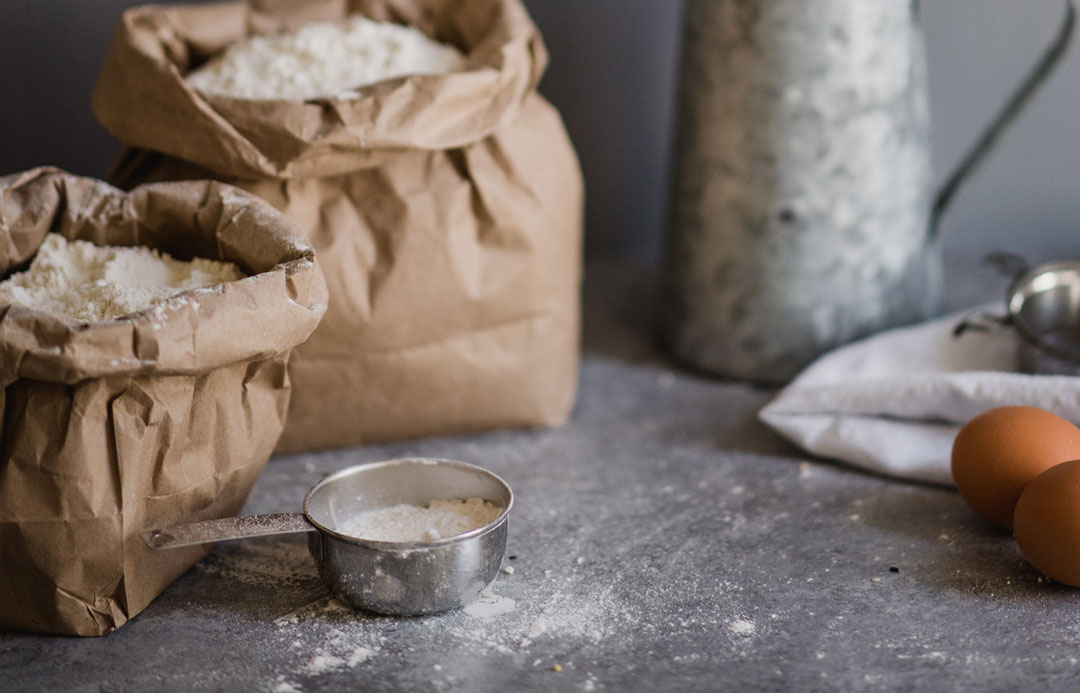 bakery-43 A rustic food photography scene featuring baking ingredients on a dark wooden table. Brown eggs, a bowl of crumbled cheese, golden raisins, sugar, flour, and baking powder are arranged on a textured gray cloth, alongside vintage spoons and a rolling pin.